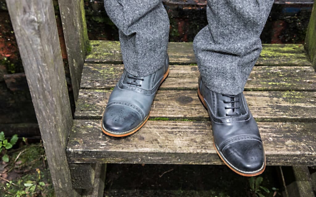 Close‑up of grey leather brogue‑style dress shoes paired with textured grey tweed trousers, photographed on a weathered wooden step in an outdoor rustic setting