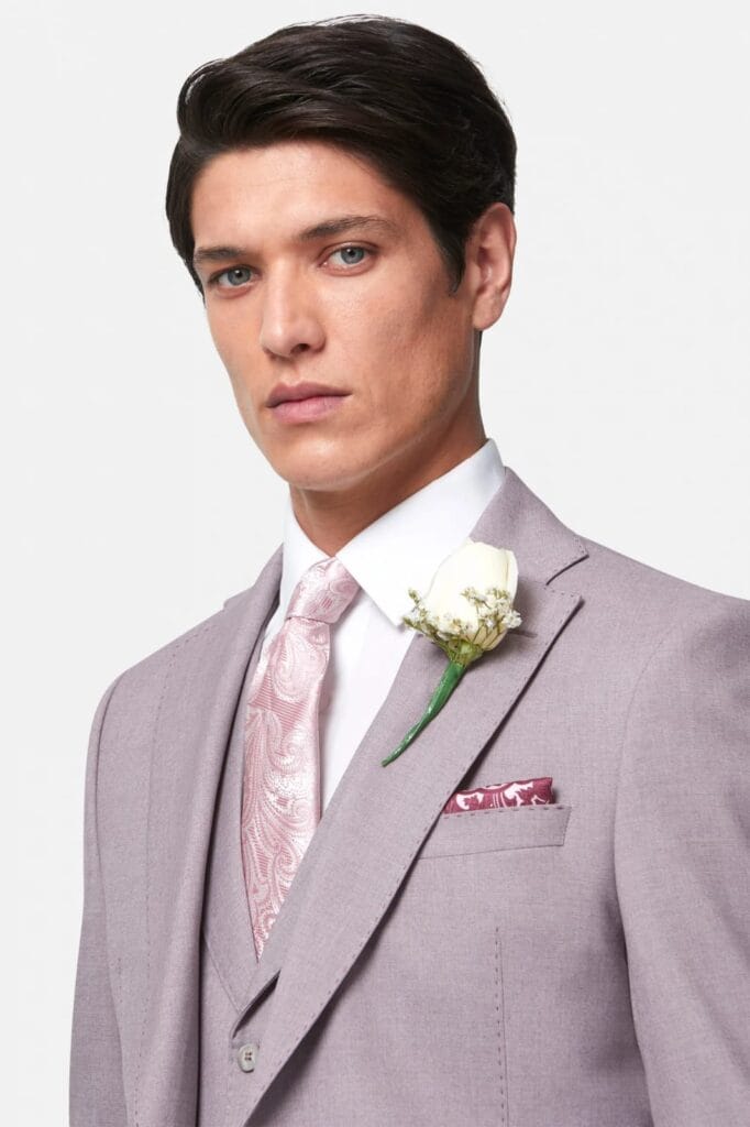 Groom wearing a light mauve three‑piece wedding suit with a paisley pink tie, white shirt, and a white rose boutonnière, styled with a coordinating patterned pocket square.