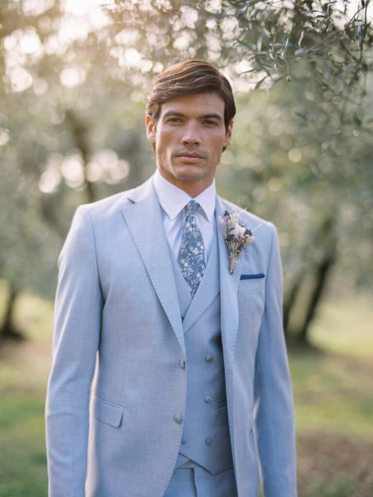 Groom wearing a light blue three‑piece wedding suit with a patterned tie and a rustic dried‑flower boutonnière, standing outdoors among softly lit trees.